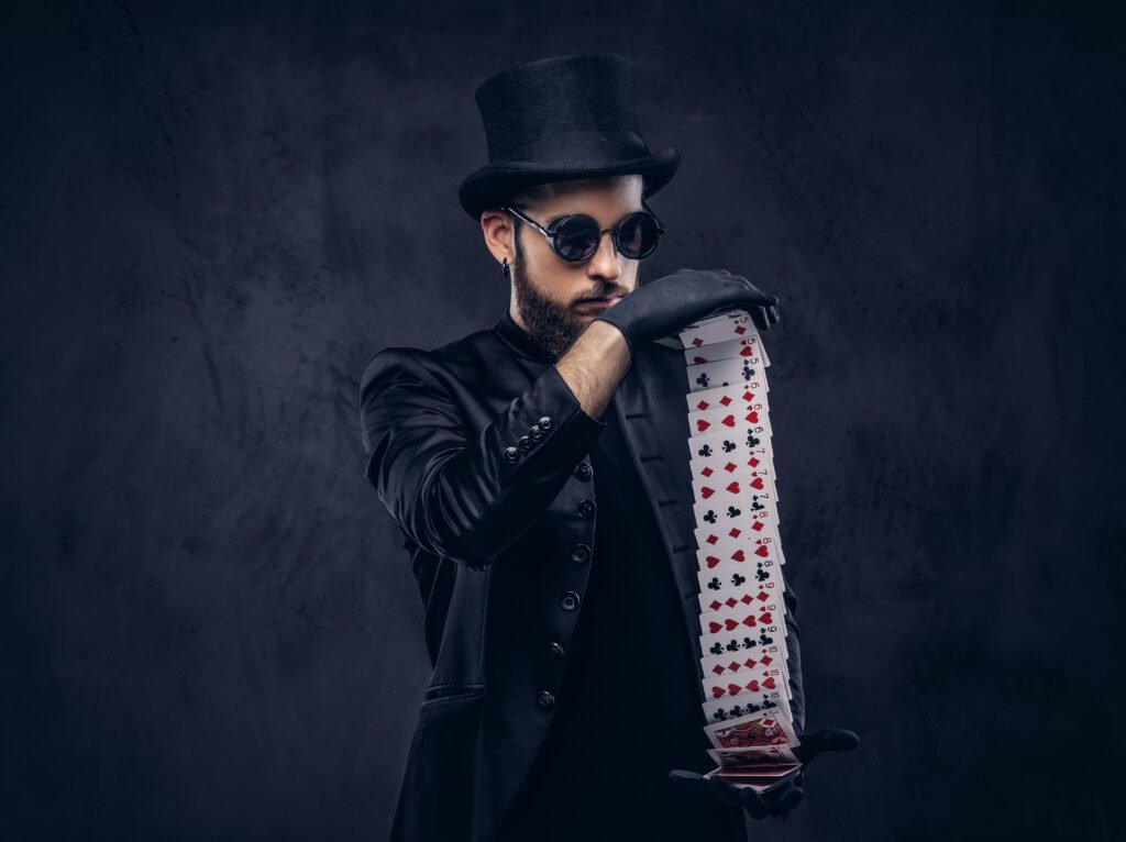 Magician in a black suit, sunglasses and top hat, showing trick with playing cards on a dark background.